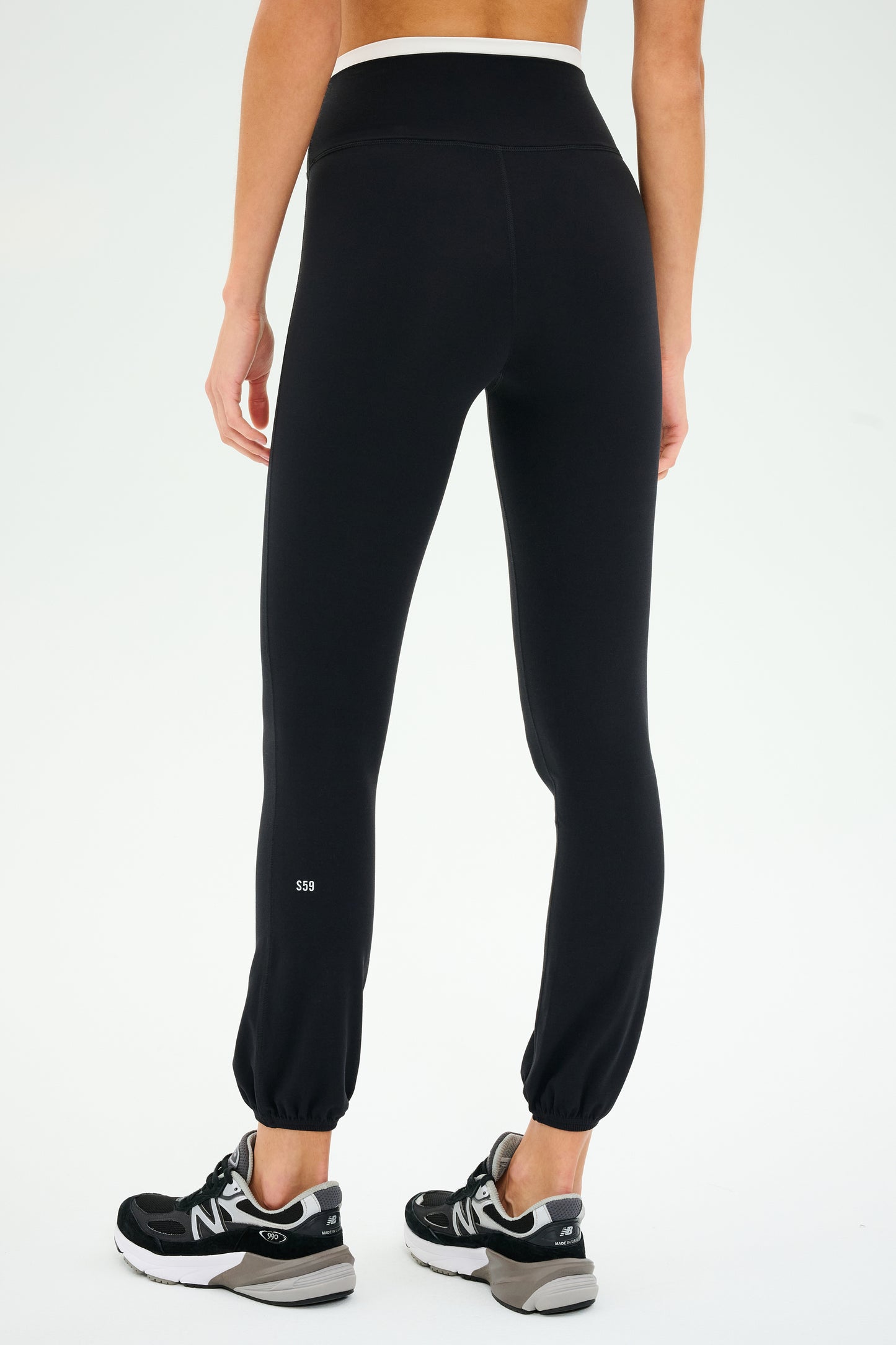 A person showcases SPLITS59's Icon Dual High Waist Airweight Leggings in black, paired with sleek black sneakers, against a simple backdrop. The luxe fabric of these leggings ensures comfort and style, ideal for any occasion.