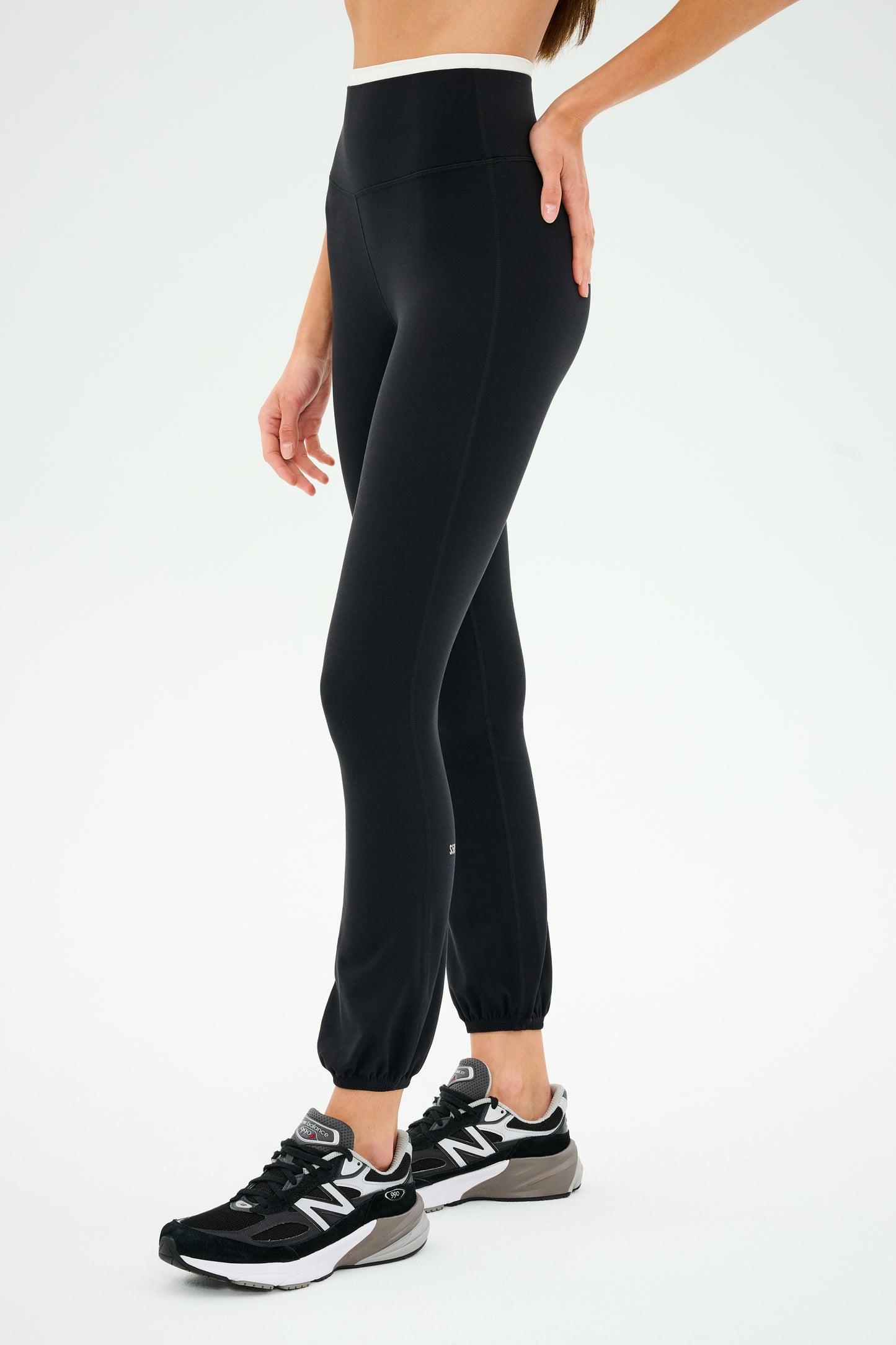 The person is confidently standing on a white background wearing SPLITS59's Icon Dual High Waist Airweight Legging in black with black sports shoes.