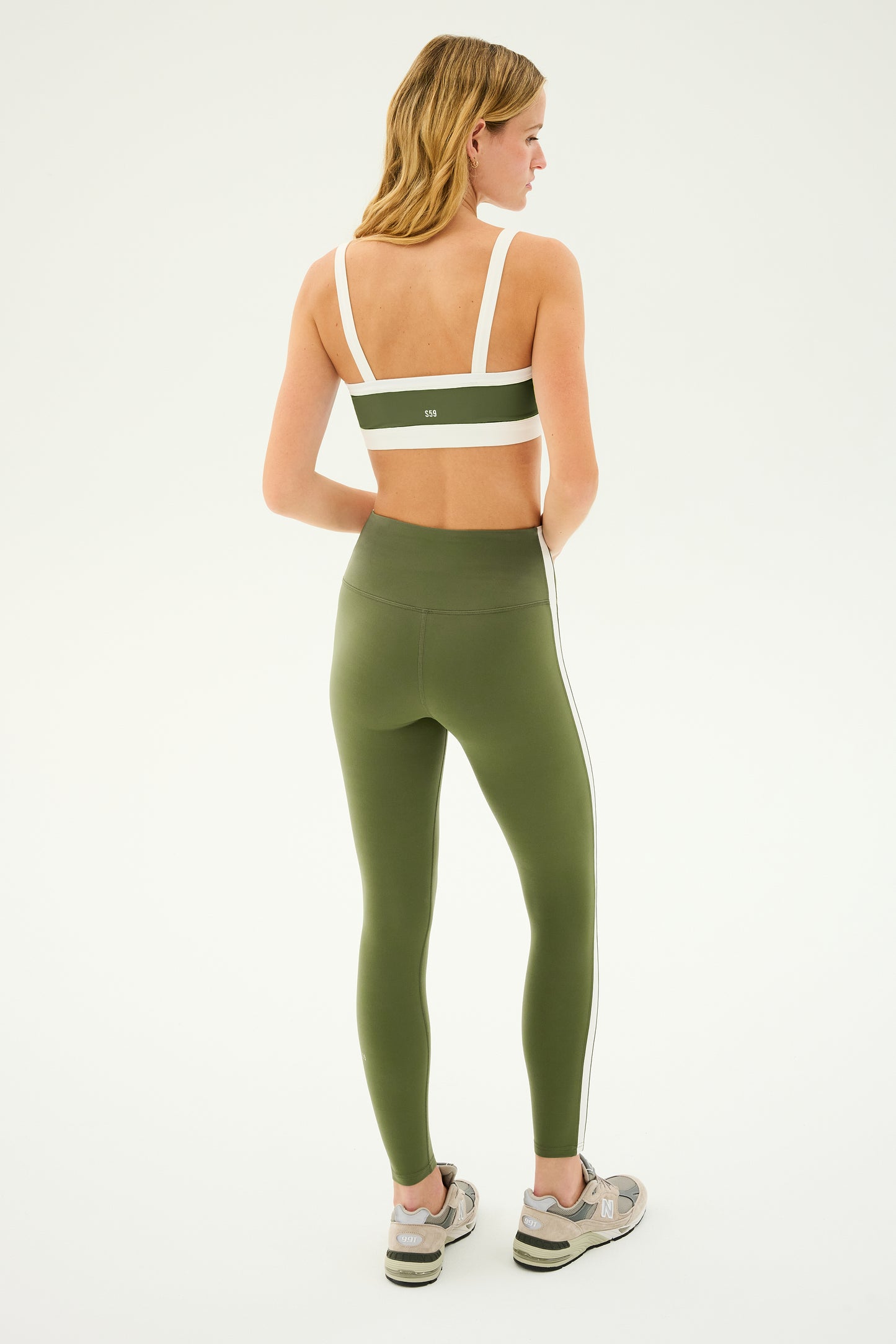 A person with blonde hair is standing turned away from the camera, wearing a Monah Rigor Bra in Olive/White by SPLITS59, matching high-waisted leggings with white stripes on the sides, and gray sneakers.