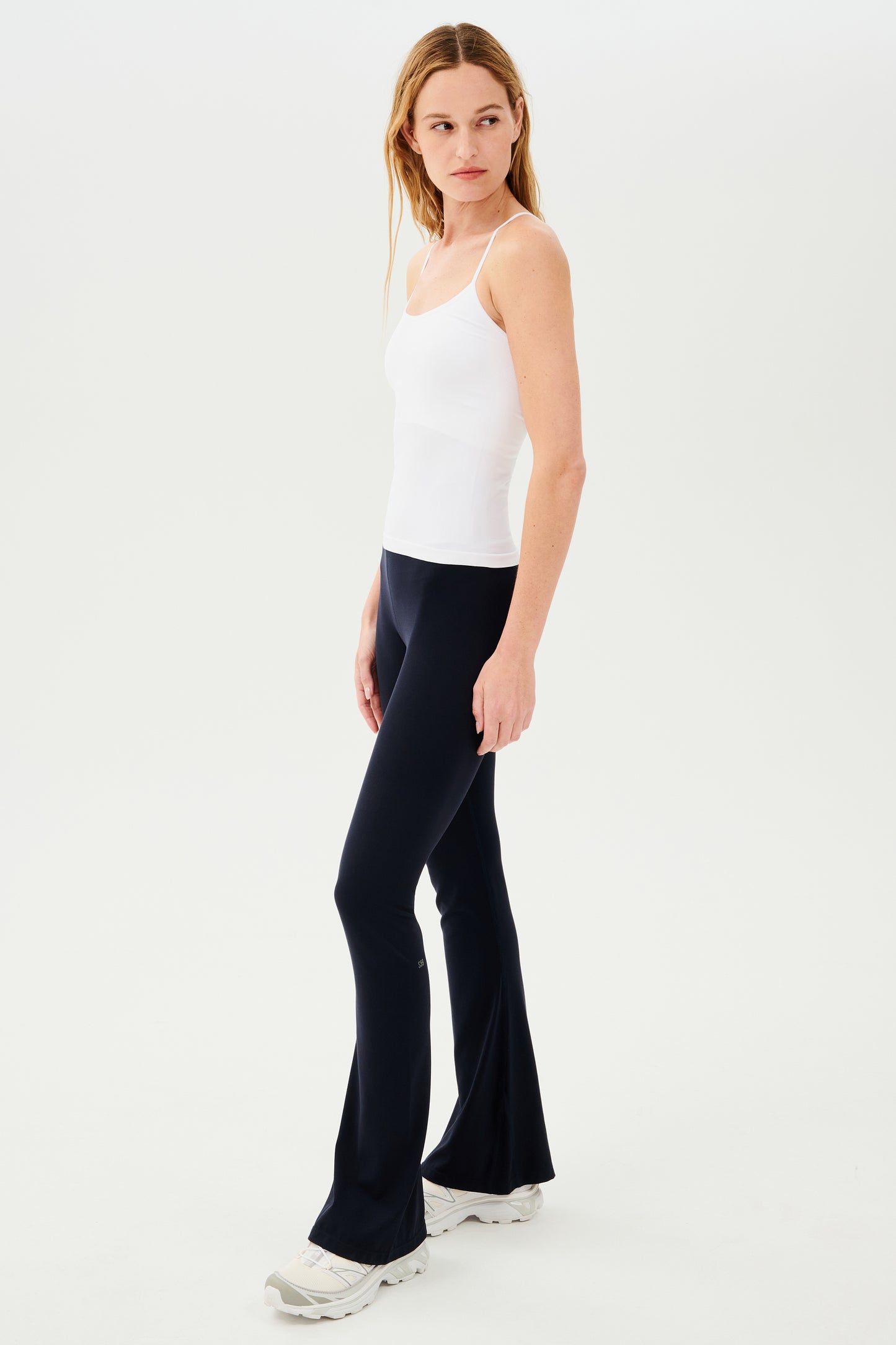 A woman wearing black high-waist flared leggings made of SPLITS59's Raquel High Waist Airweight Flare - Indigo fabric and a white tank top.