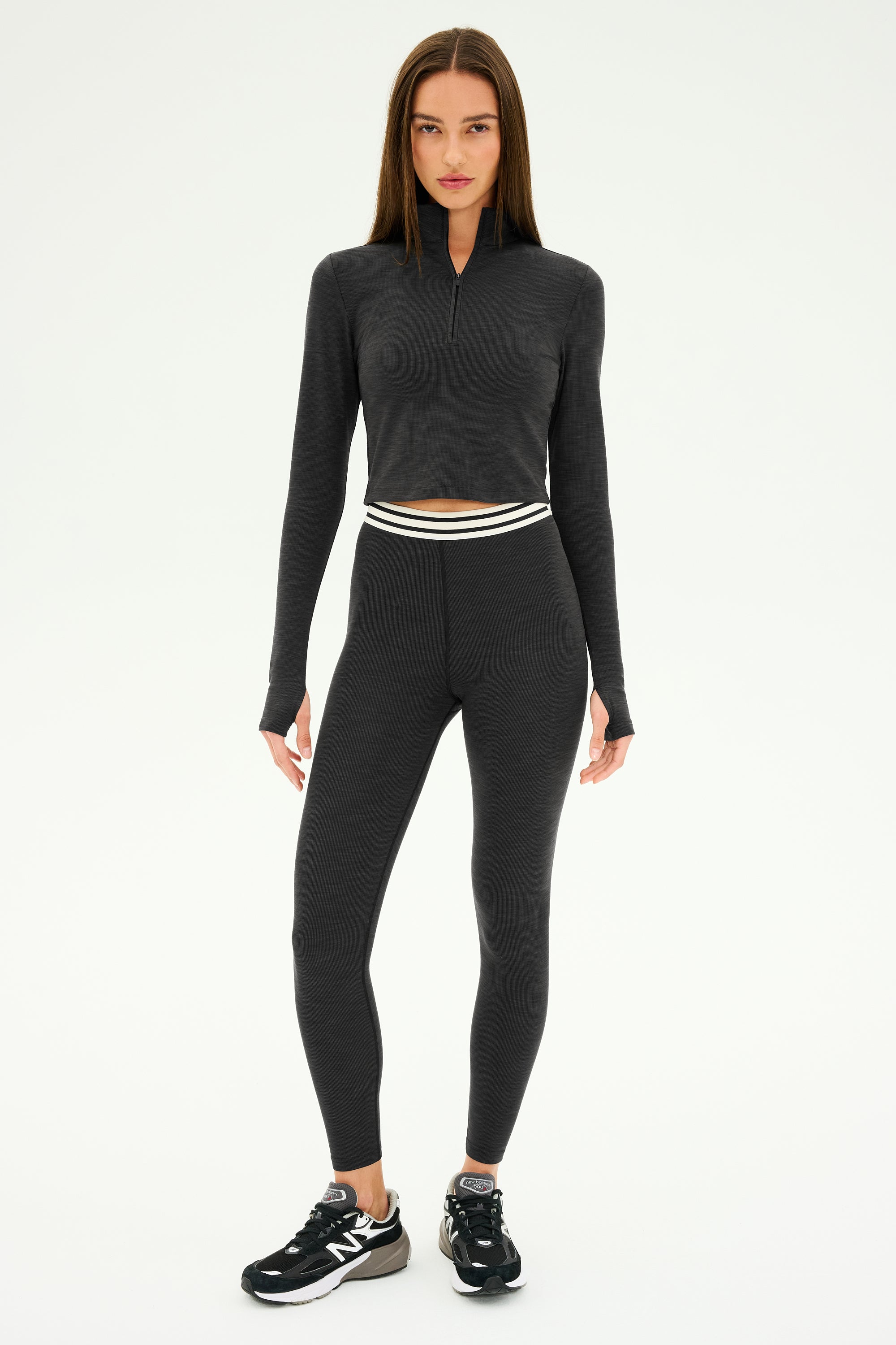 A person wearing a stylish athletic outfit from SPLITS59, featuring the Stevie Active Rib Half Zip in Heather Graphite and leggings crafted from active rib fabric, pairs with black and white sneakers. They stand confidently against a white background, ready for gym workouts.