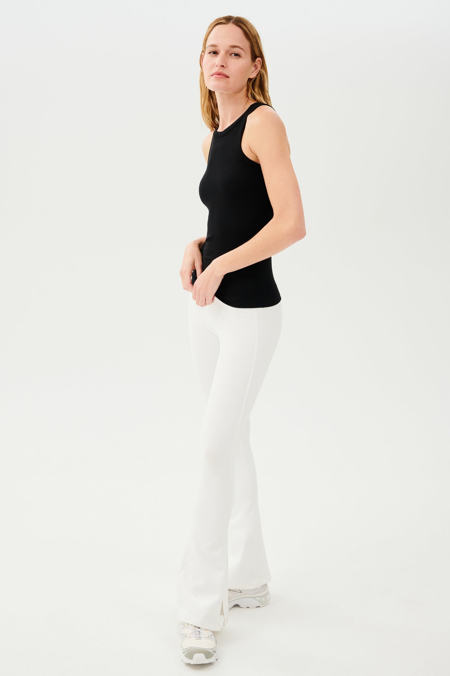 Full front side view of woman with blonde straight hair wearing white high waist below ankle length legging with wide flared bottoms with split hem flare opening and black racerback tank top. Paired with white and gray shoes.