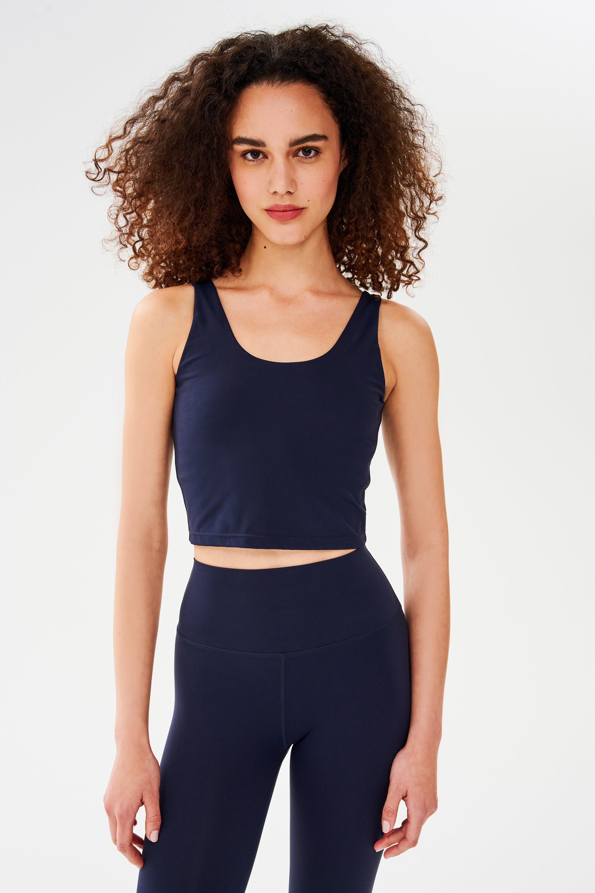 Front view of woman with curly dark brown hair wearing a dark blue bra crop tank with dark blue leggings 