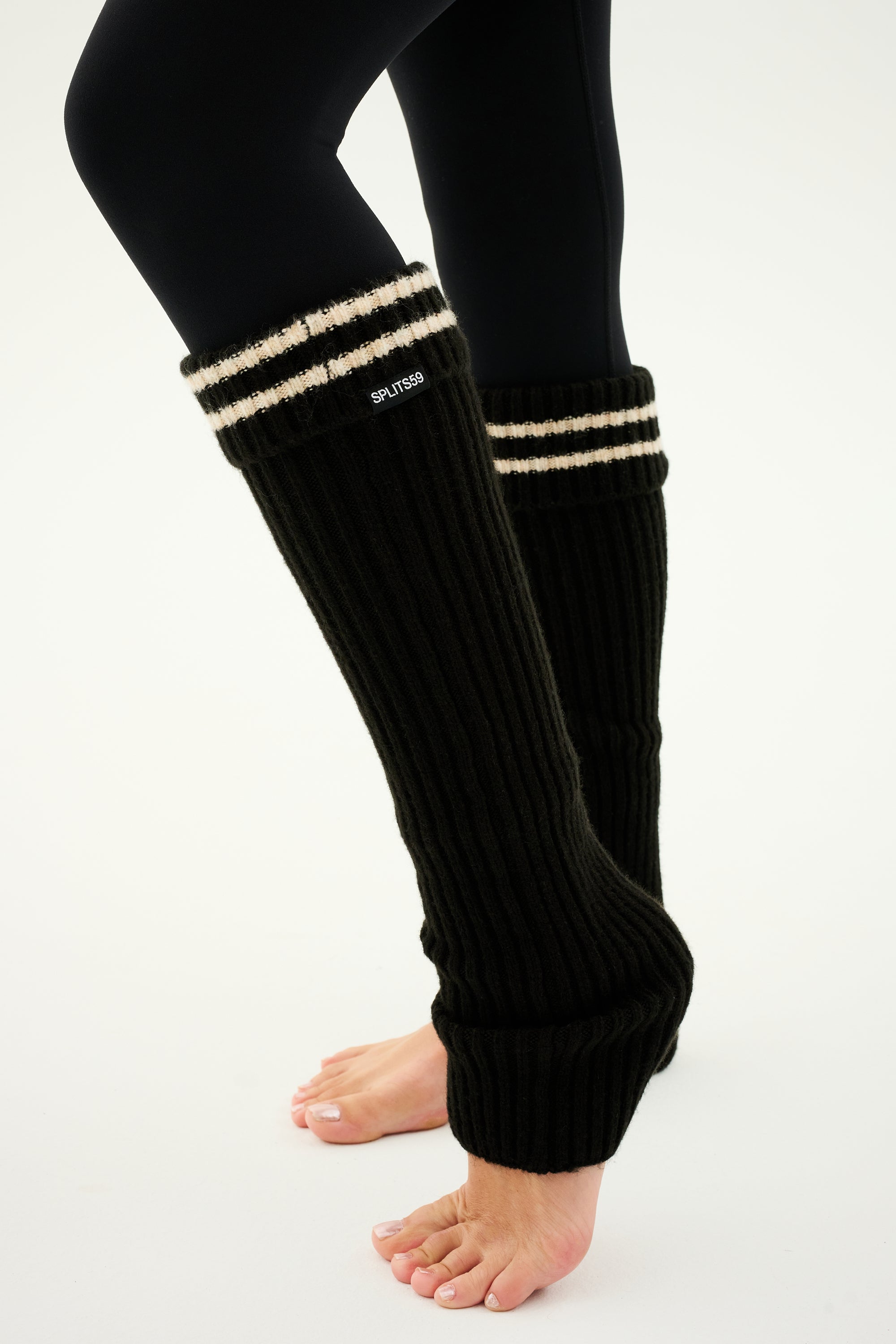 A person wearing SPLITS59 Leg Warmer - Black/Creme over black leggings, standing barefoot on a plain background.