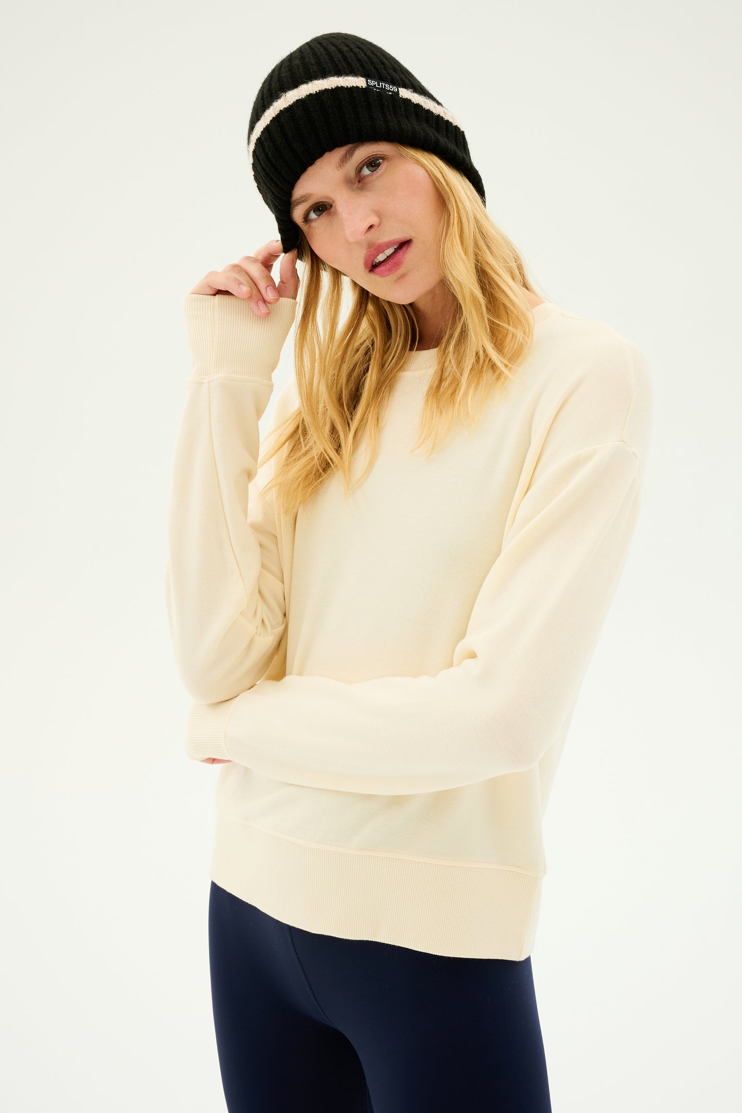 A person stands against a plain background wearing a light beige sweatshirt, dark pants, and the cozy SPLITS59 Rib Beanie in Black/Creme, perfect for cold weather workouts.