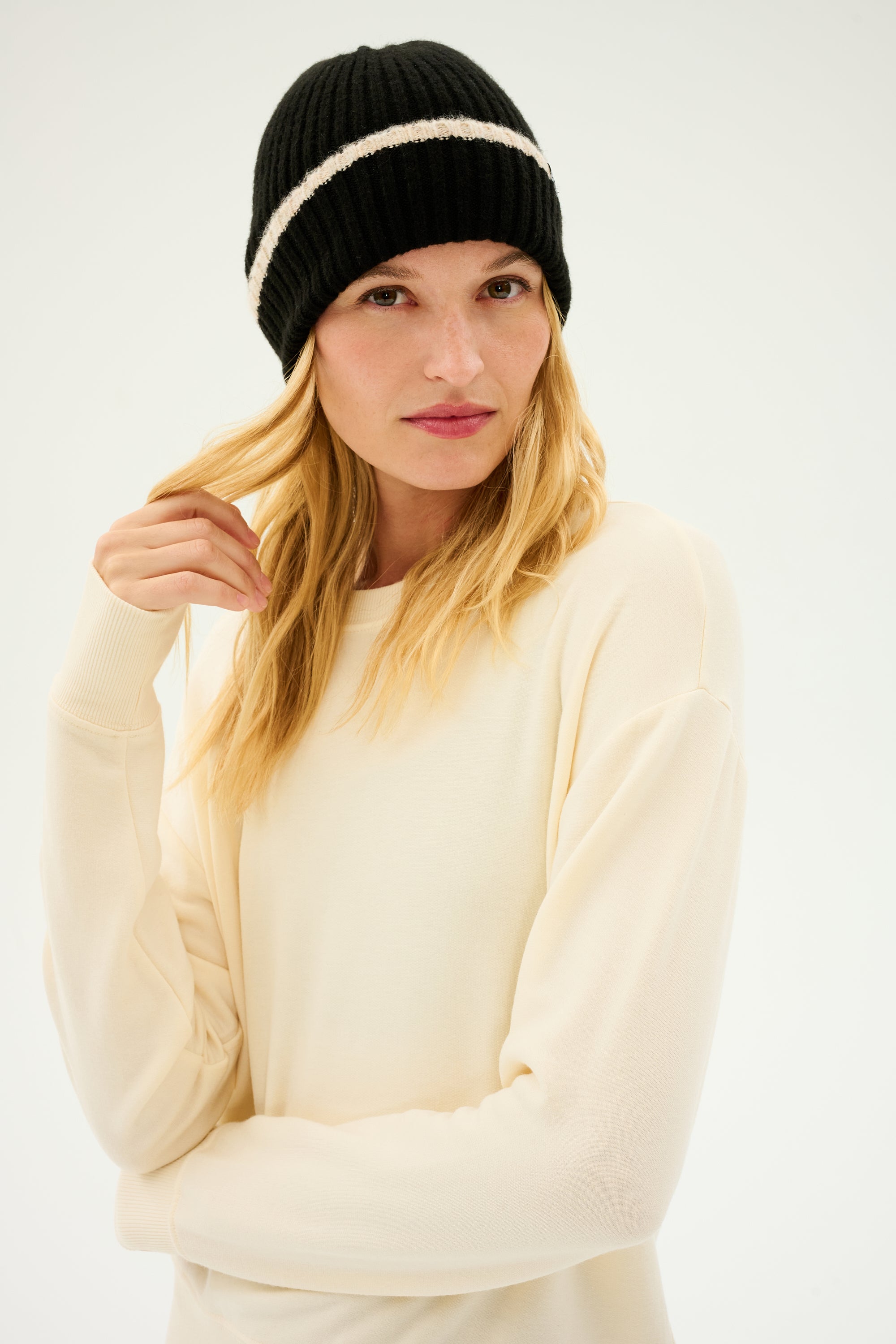 A person with long blonde hair, wearing the SPLITS59 Rib Beanie in Black/Creme and a cozy cream-colored sweater, looks directly at the camera.